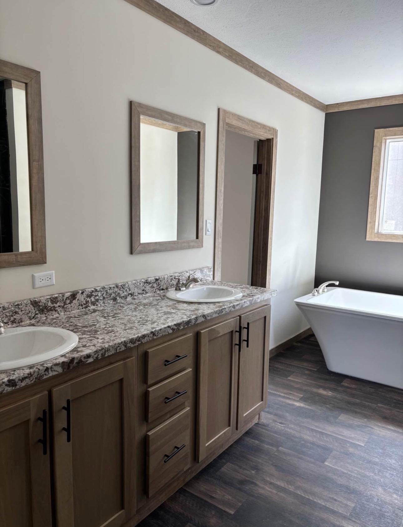 A modern bathroom with dual sinks set in a marble countertop, wooden cabinets, two mirrors, and a freestanding bathtub. Warm, inviting tones are present.