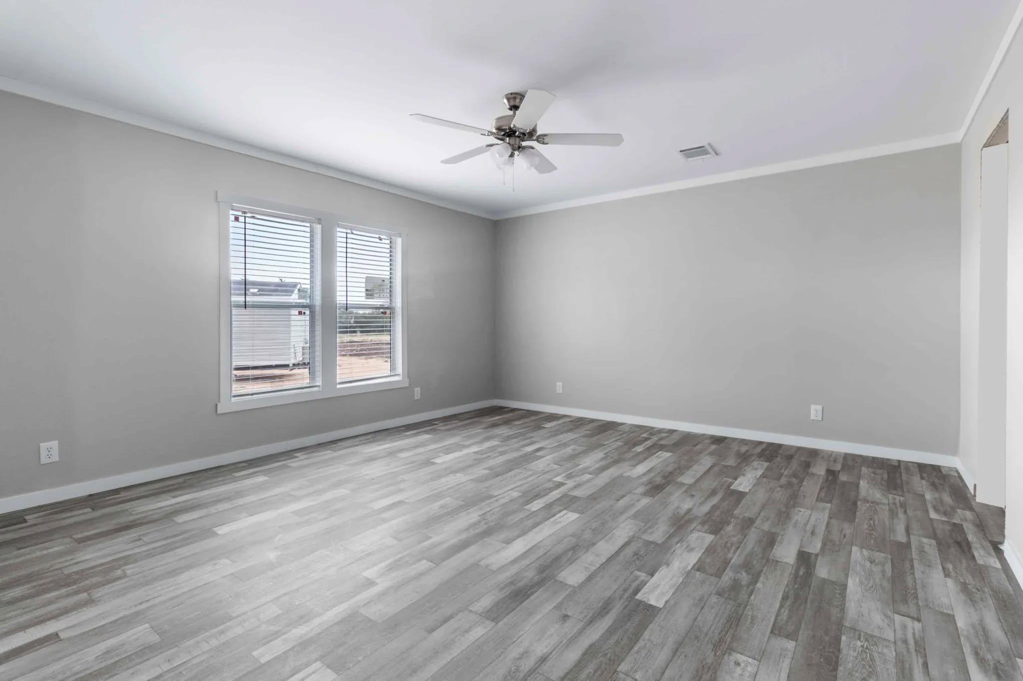 A bright, empty room with light gray walls, a ceiling fan, and wood-patterned flooring. Sunlight streams through two windows, creating a serene atmosphere.