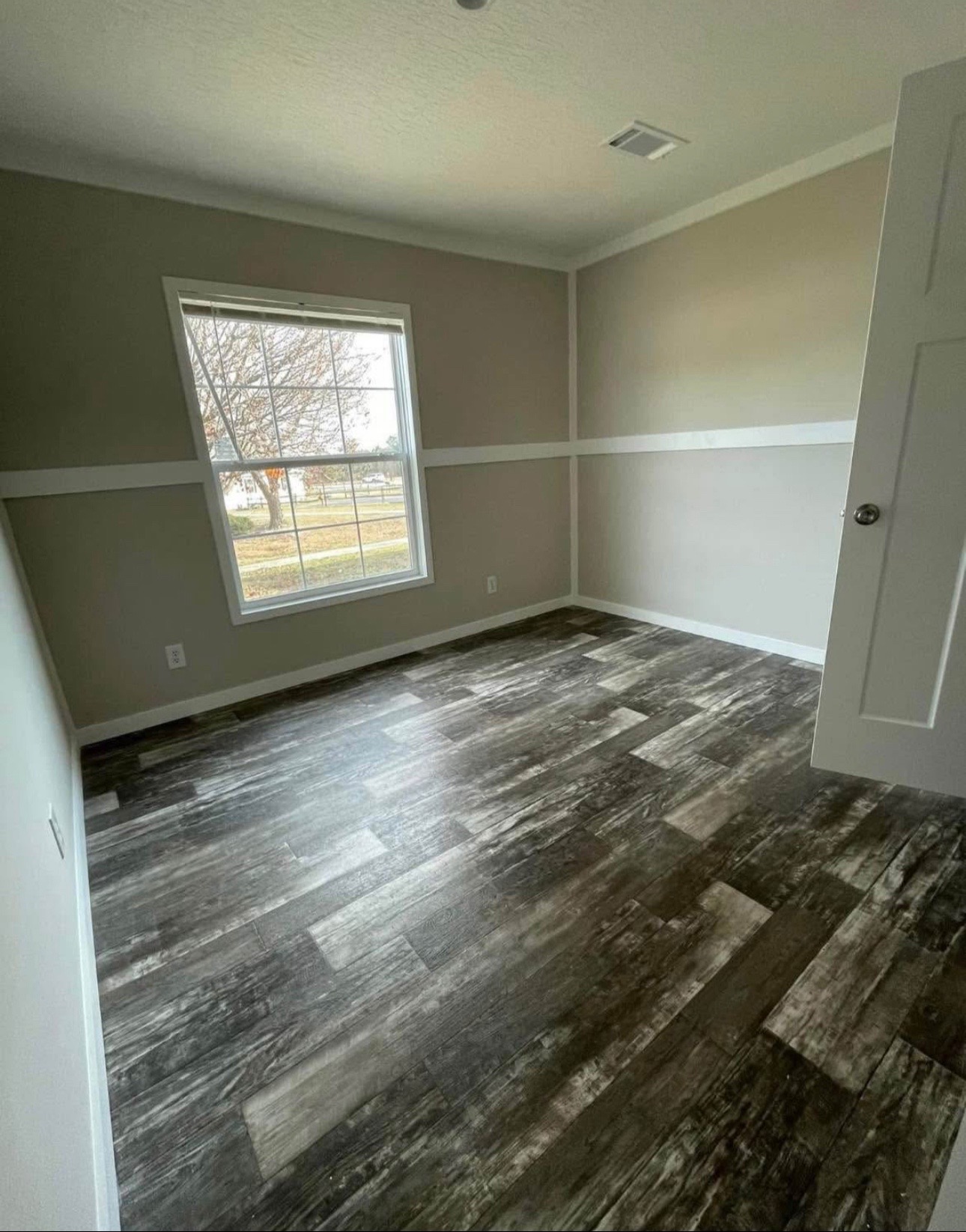 Empty room with light gray walls and dark wood-textured flooring. Large window with a view of trees and grass. Open door. Calm and minimalist.
