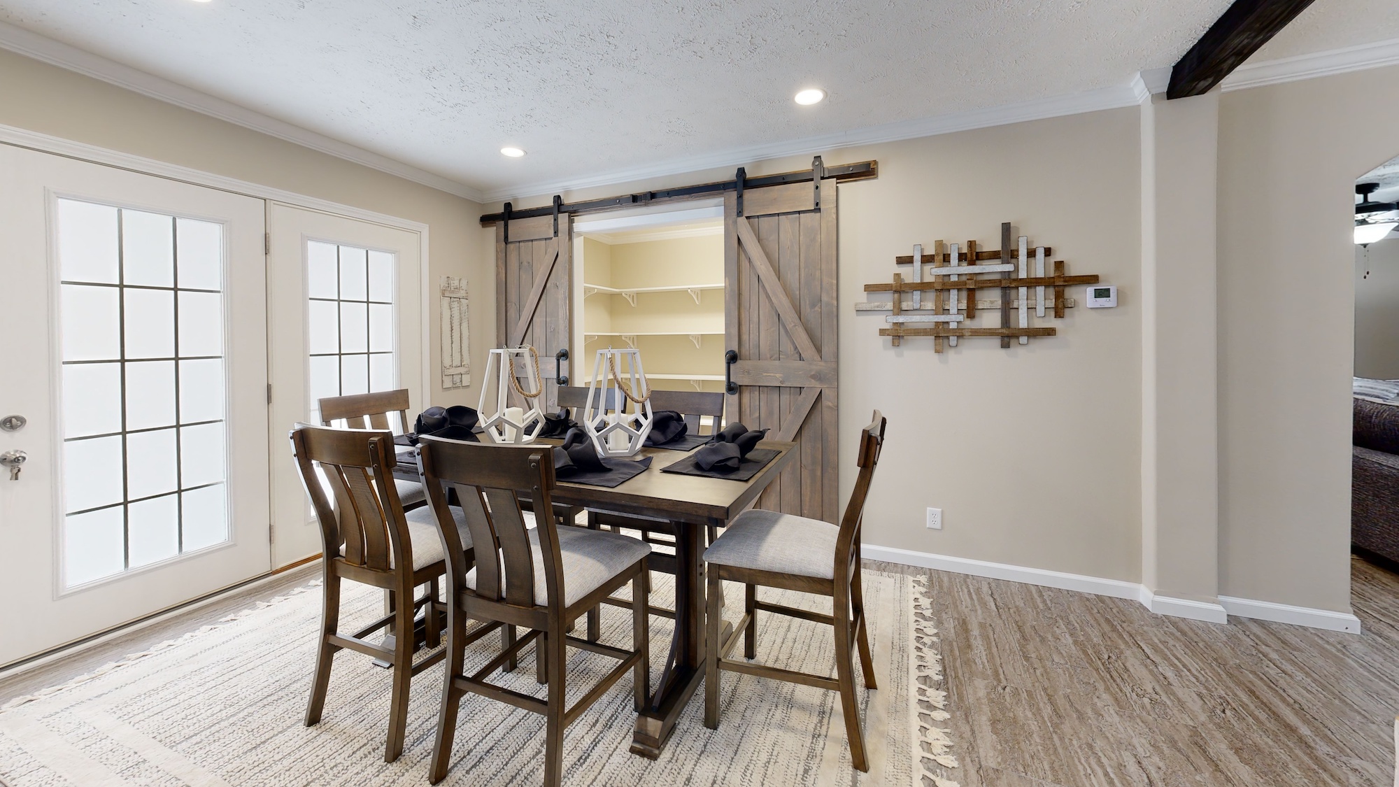 Dining room with a wooden table set for four, featuring dark napkins and modern decor. Barn doors open to a lit pantry. Warm tones and elegant ambiance.