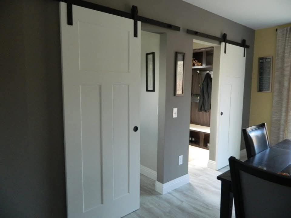A modern room with two white sliding barn doors on dark tracks, opening to a hallway with wall art. A coat and bag hang in the entryway, beside a dining table.