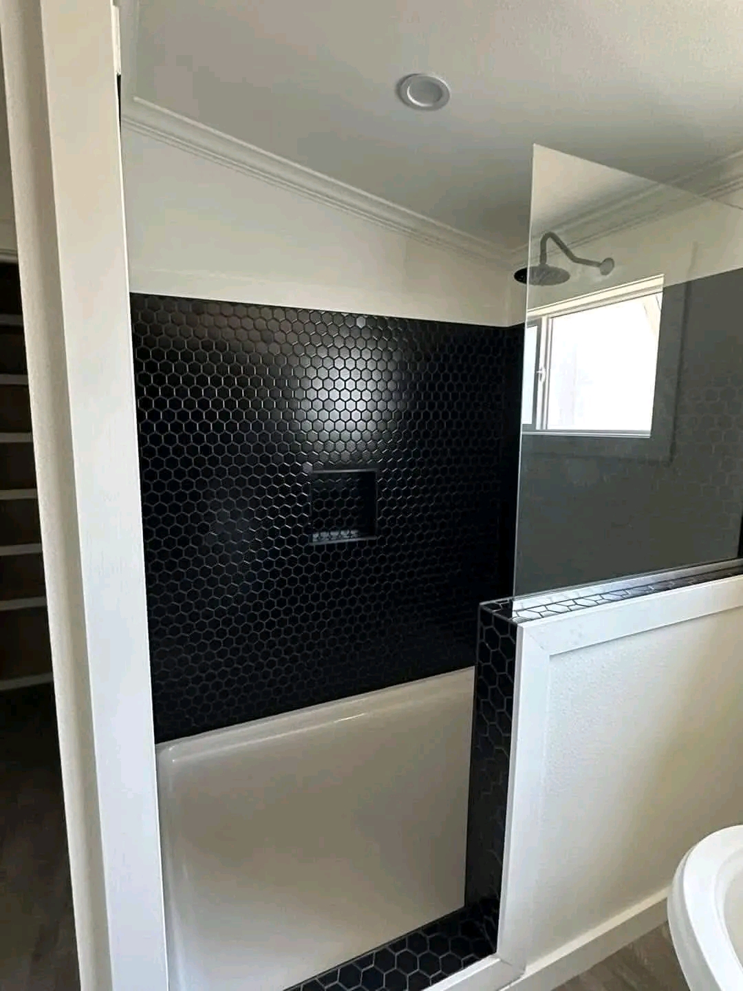 Modern bathroom featuring a white bathtub, black hexagonal tiled wall with a recessed shelf, glass shower panel, and window, creating a sleek, minimalist feel.