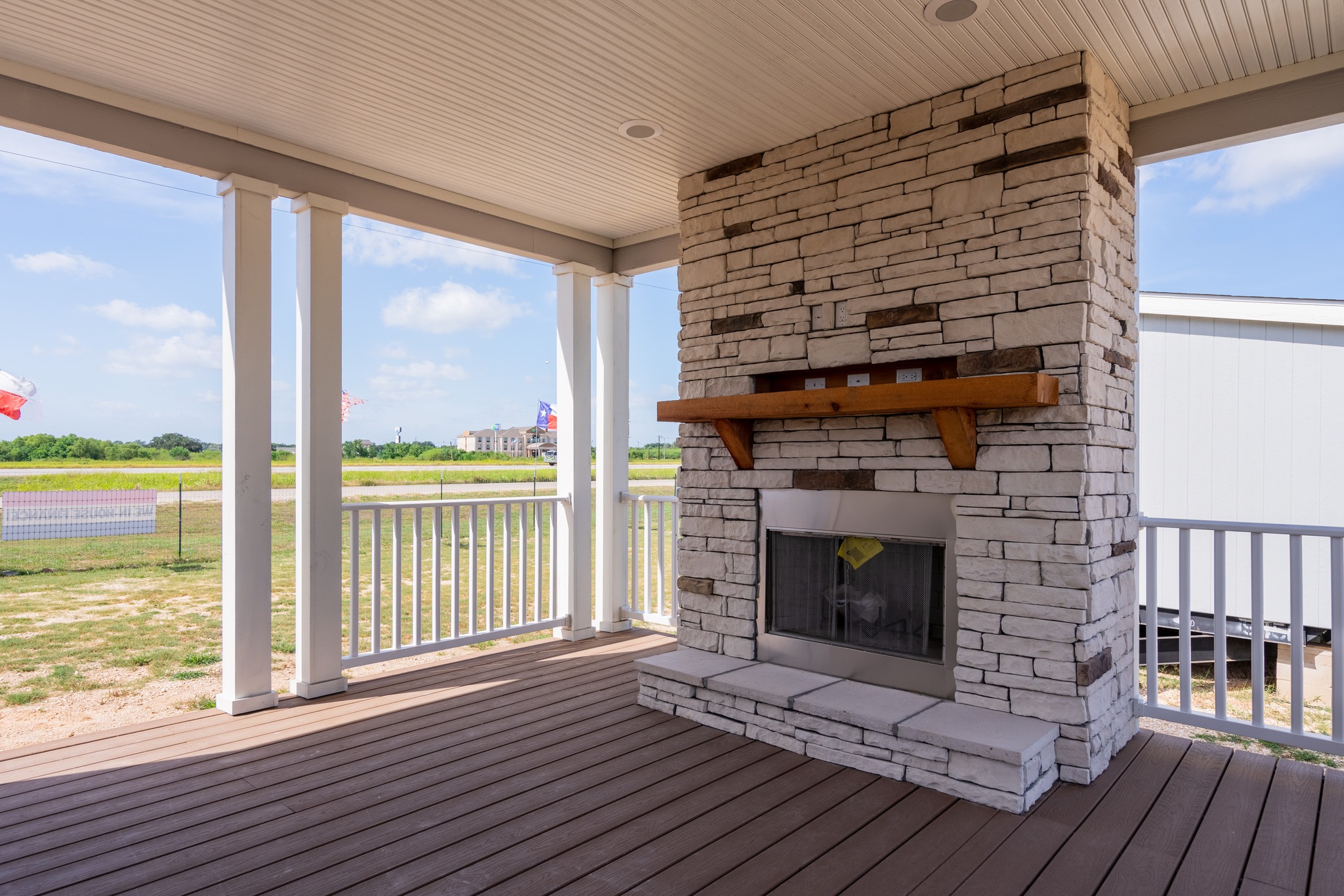 Covered porch with wooden decking and a large stone fireplace. White railing encircles the porch with a sunny, open field and blue sky beyond. Peaceful and inviting.