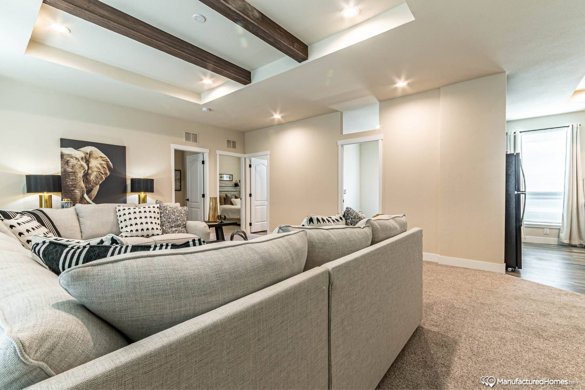 Spacious living room with large gray sectional sofa, patterned pillows, and elephant artwork. Tray ceiling with wooden beams and soft lighting convey a modern, cozy ambiance.
