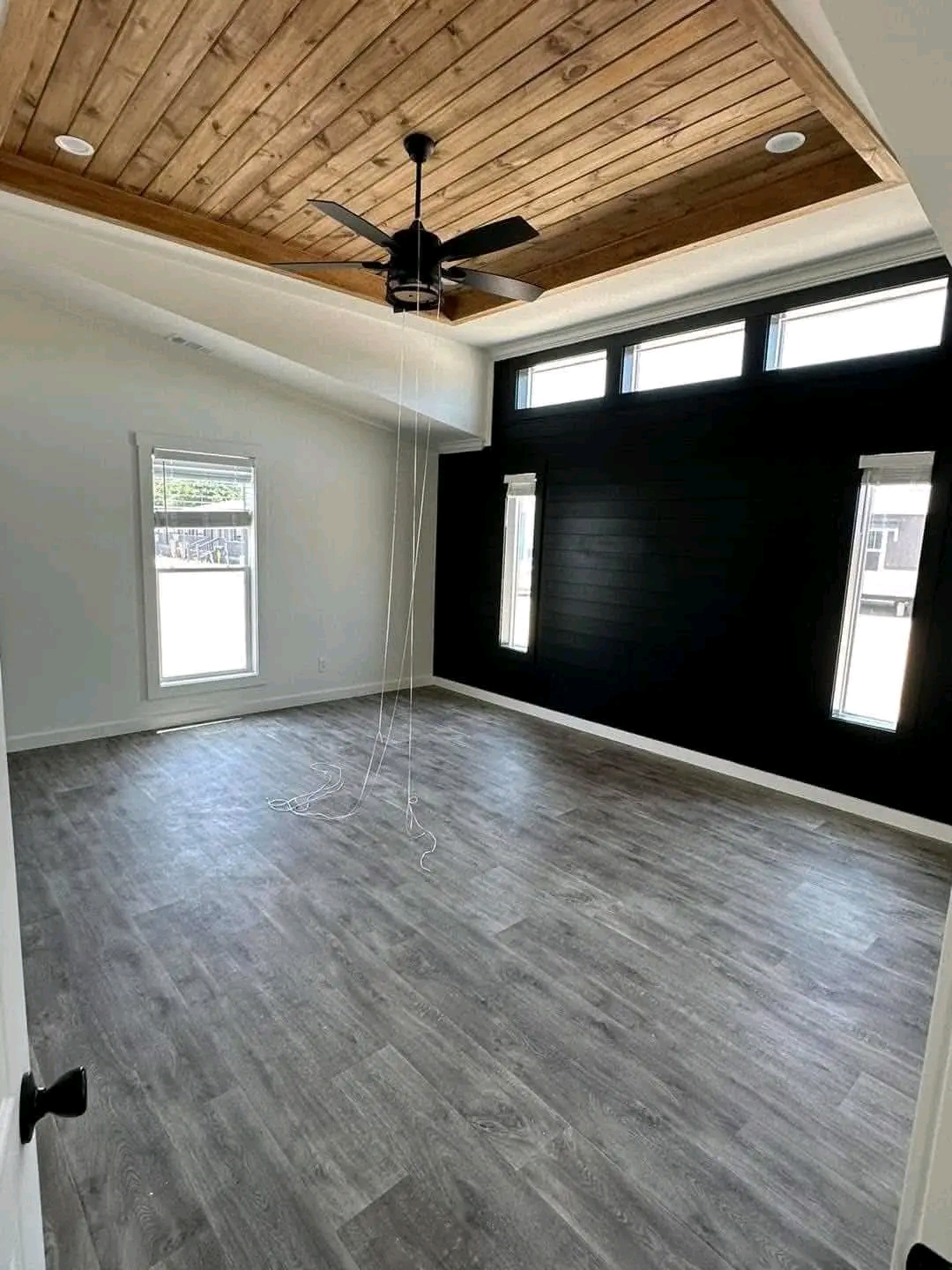 Modern room with light wooden flooring, black accent wall featuring narrow windows, and rustic wooden ceiling with a black ceiling fan. Bright and spacious.