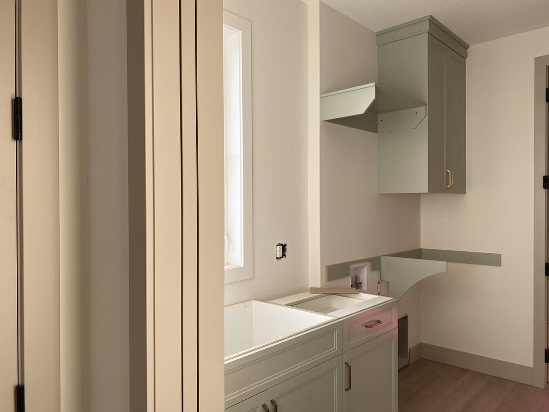 Laundry room with light green cabinets, a large sink, and wooden flooring. Soft light filters through a window, creating a calm and inviting atmosphere.