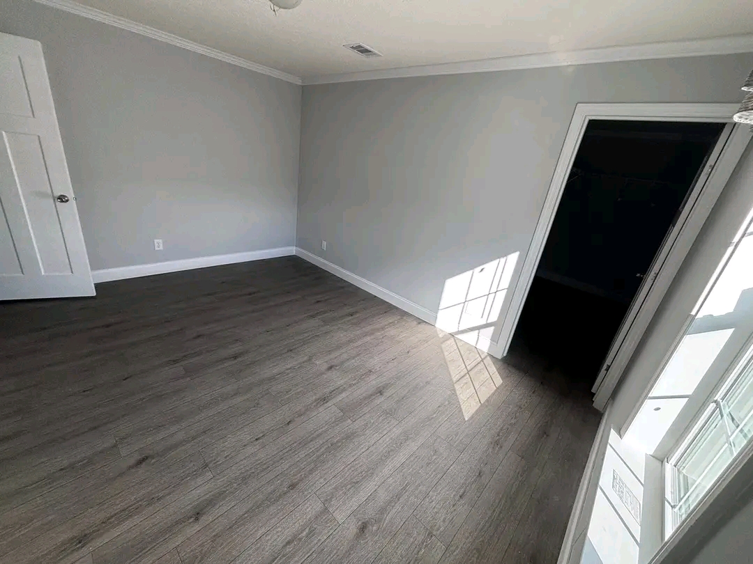 Empty room with gray walls and wood flooring. Sunlight streams through a window, casting a geometric shadow on the floor. Doorway visible on the right.