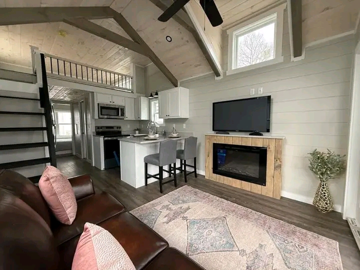 Cozy living room with a brown leather couch and pink pillows. Features a TV above a fireplace, modern kitchen with gray stools, and a rustic loft.