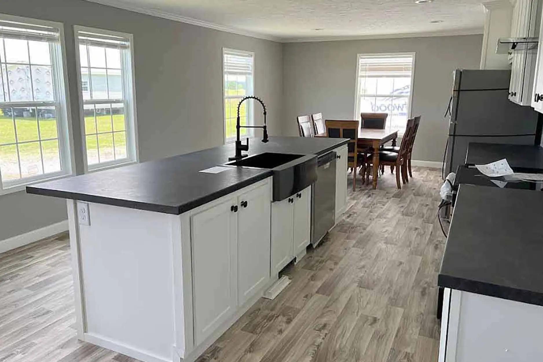 Modern kitchen with light gray walls and wood flooring. Features a central island, black countertops, stainless steel sink, and appliances. A dining table with chairs is visible near large windows, creating a bright and airy atmosphere.