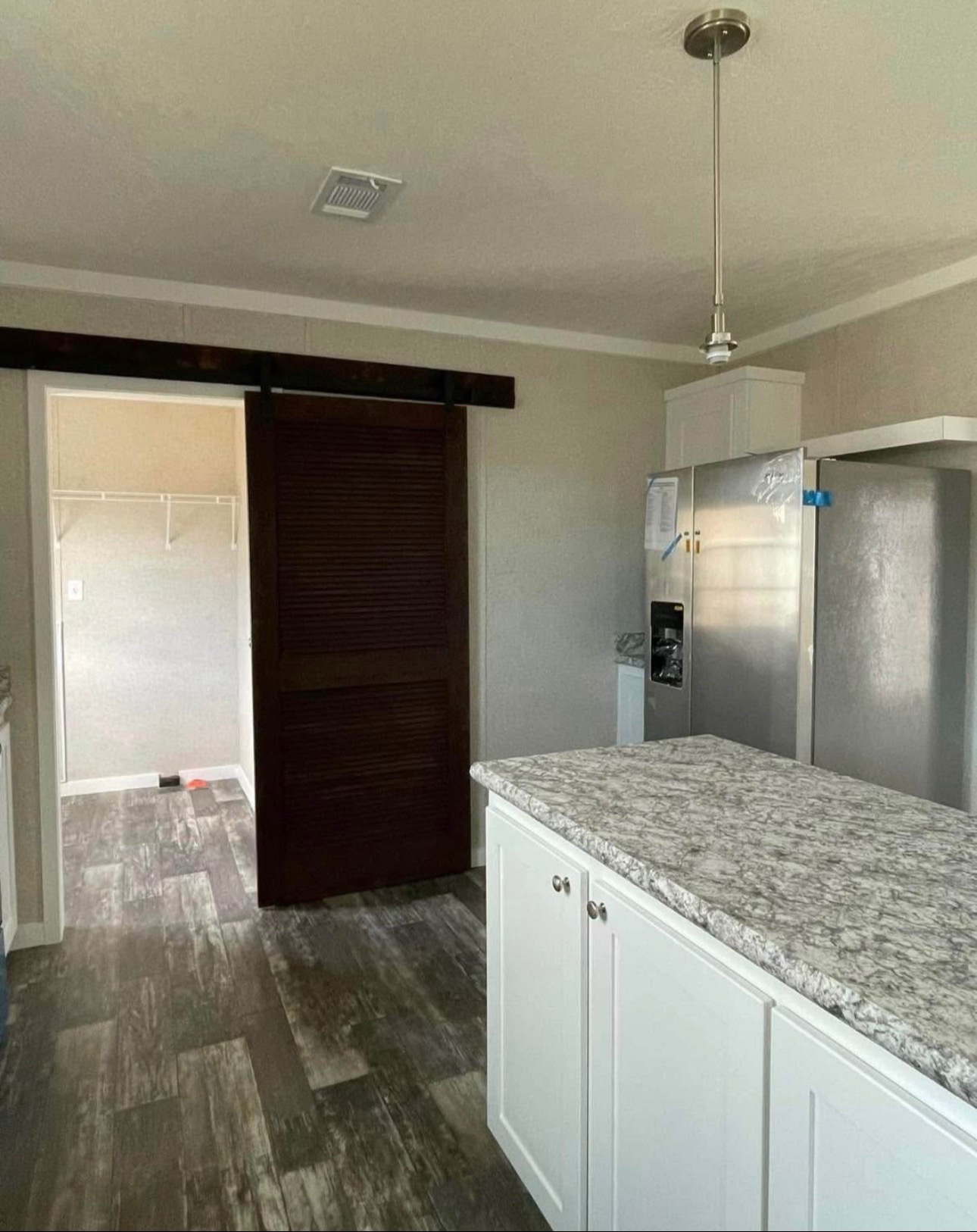 A modern kitchen with a granite island, white cabinets, stainless steel fridge, and a dark sliding pantry door. A pendant light hangs above.
