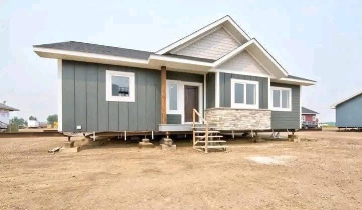 A single-story prefab house on a dirt lot. The home has gray siding, a small staircase leading to the front door, and white-framed windows. The sky is clear.