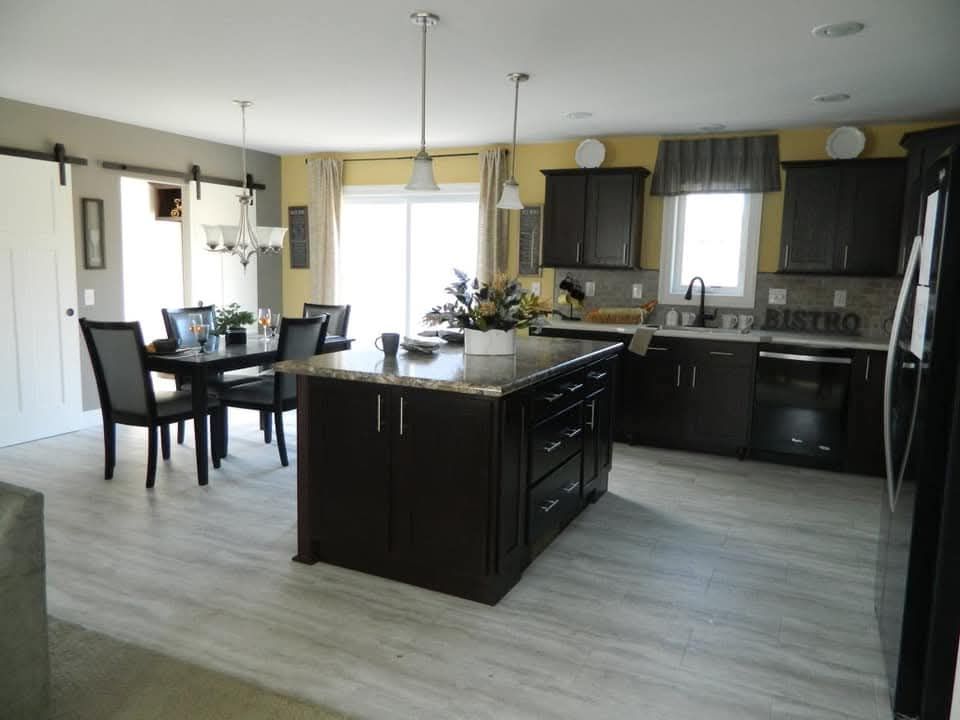 Modern kitchen with dark wood cabinets, central island, and granite countertops. Bright dining area on left, pendant lights, and light wood floors.