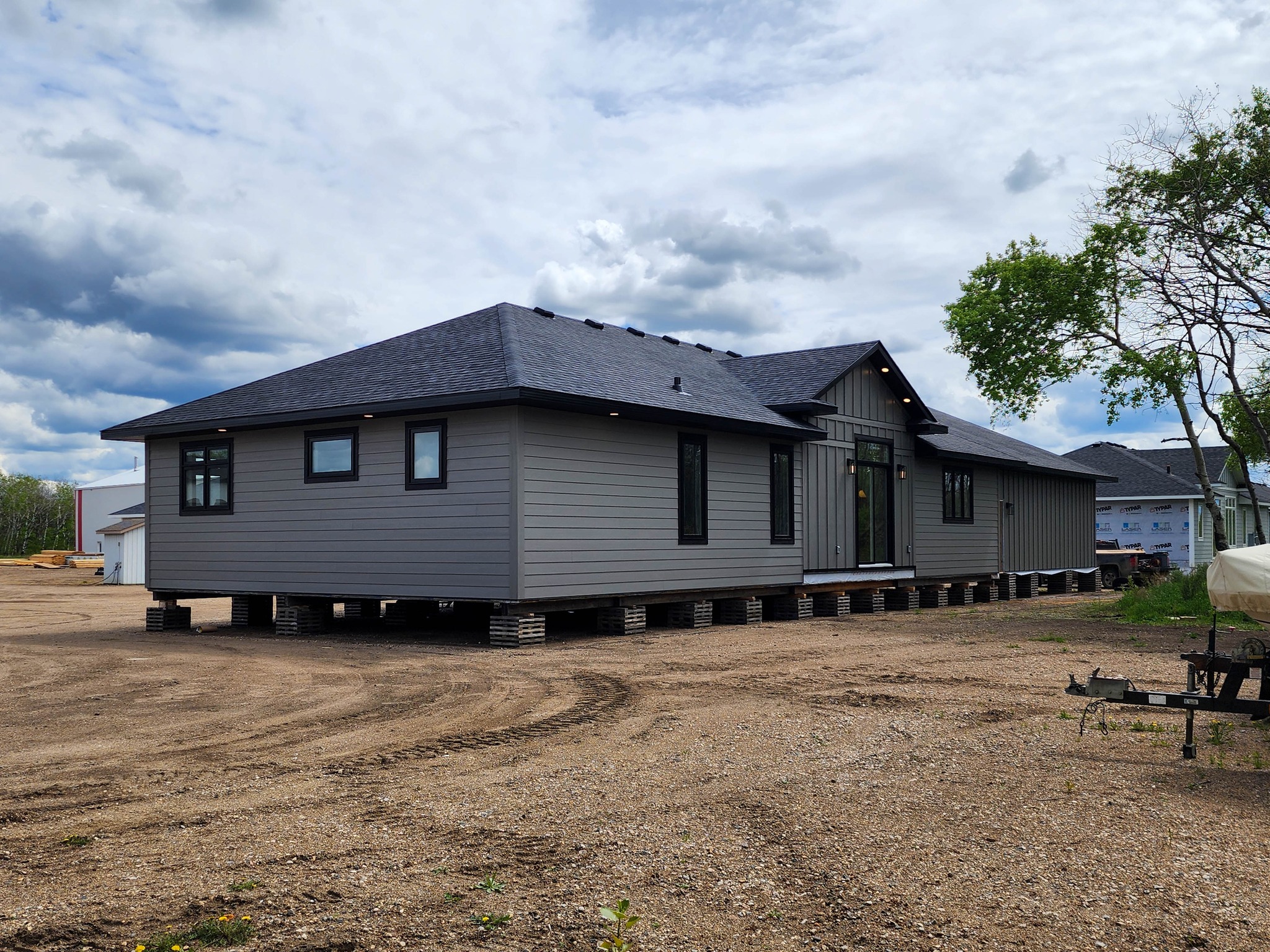 A modern, single-story grey house rests on temporary supports in a dirt lot, under a cloudy sky. Nearby, a tree adds greenery, creating a transitional mood.