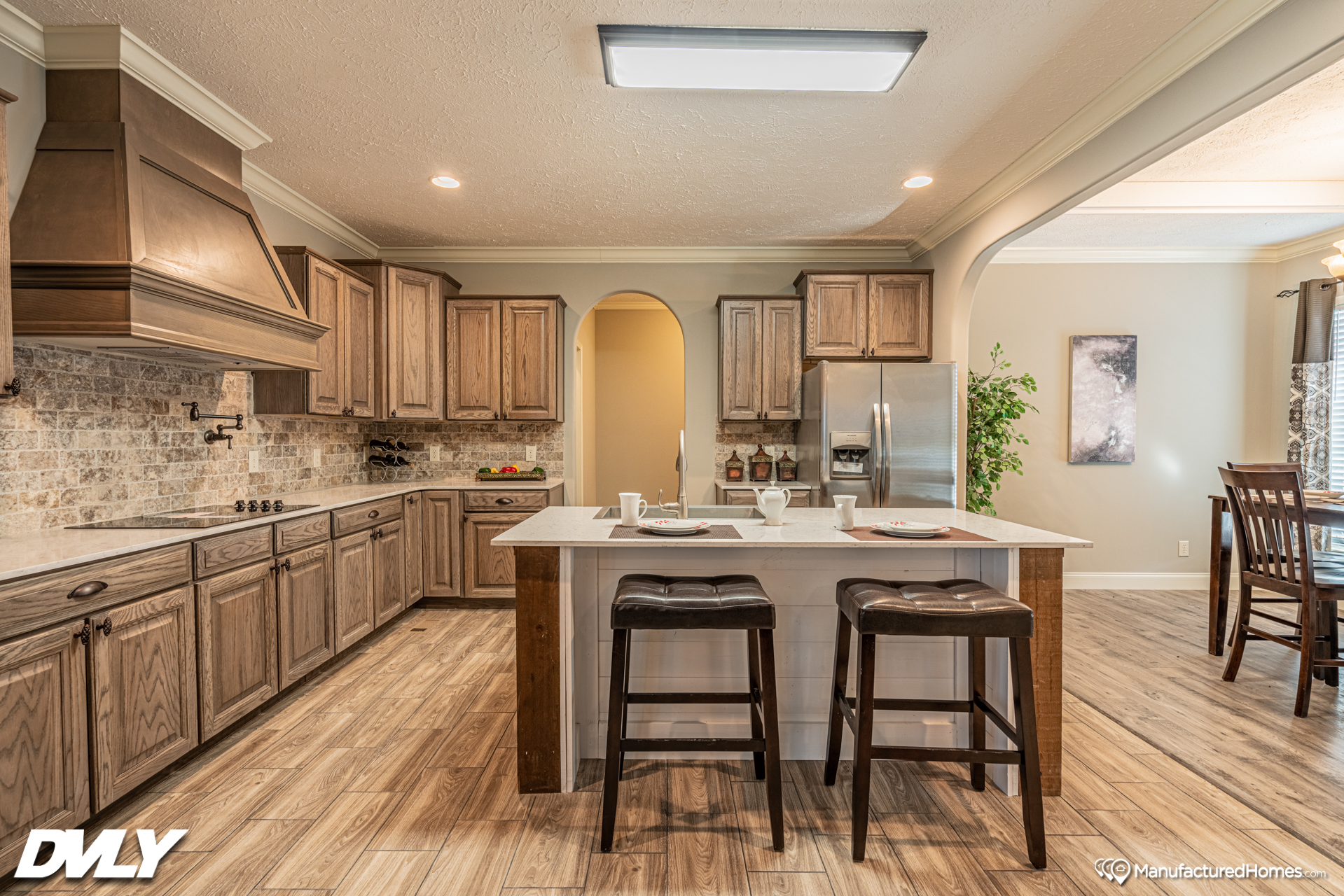 A spacious kitchen with wooden cabinets and flooring, featuring a central island with two stools, pendant lighting, and a dining area to the right.