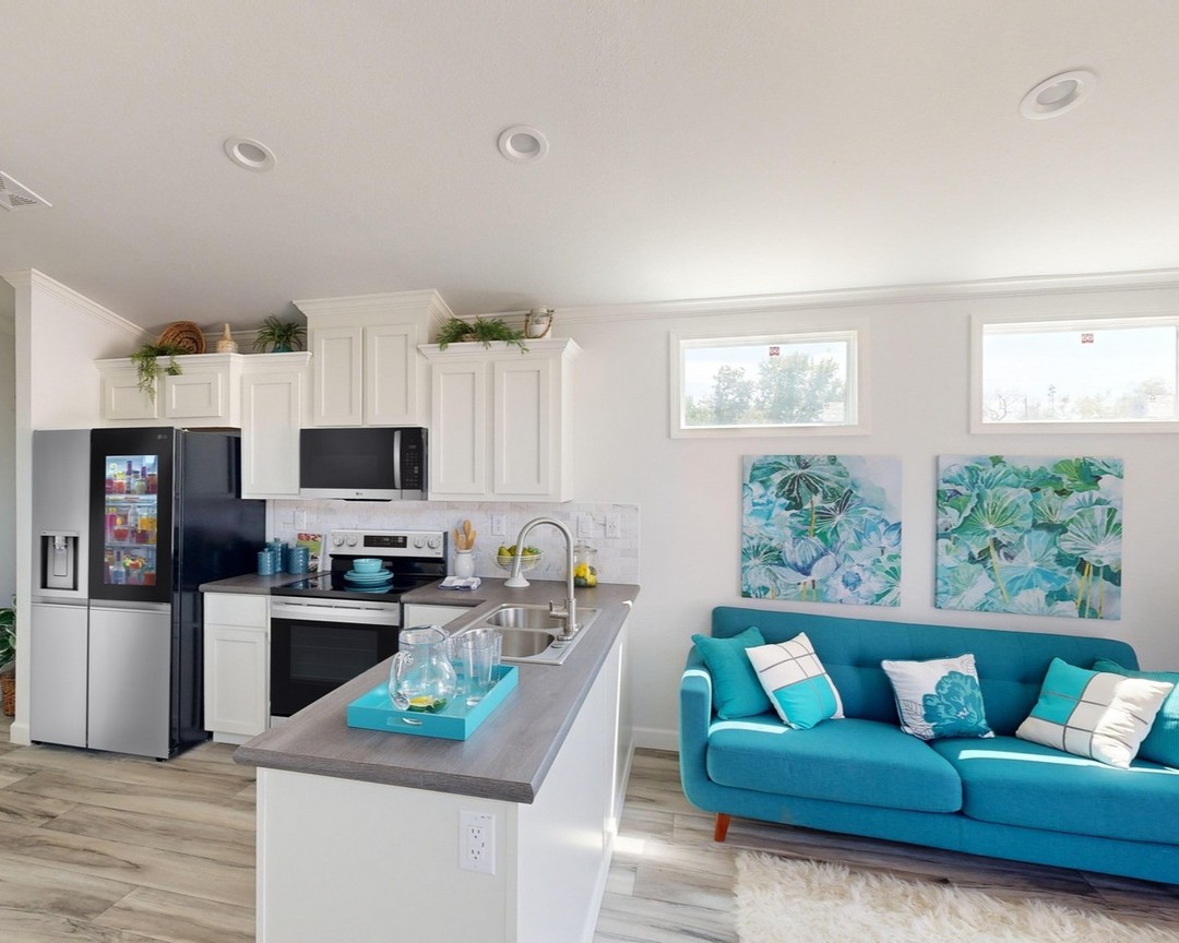 Bright, modern kitchen with white cabinets, stainless appliances, and a central island. A turquoise sofa with pillows and abstract art adds vibrancy.