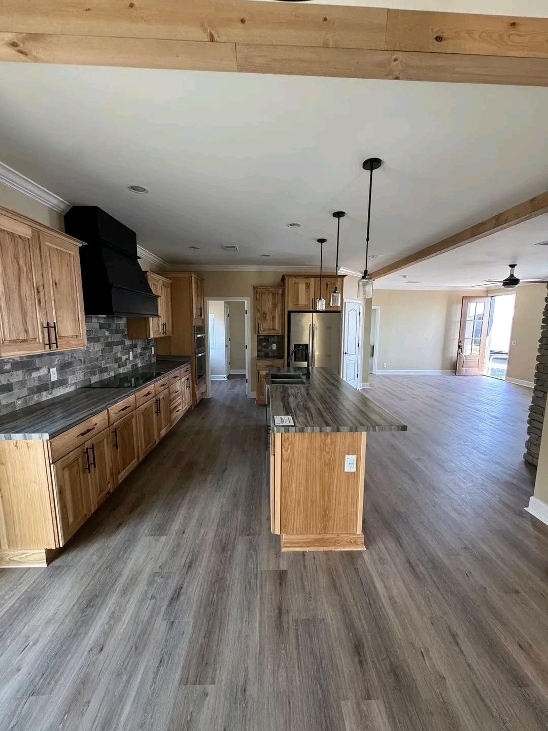 Spacious modern kitchen with light wood cabinets, gray countertops, and island. Open layout leads to a bright living area with wooden floors. Ceiling fans and stone accents add charm.