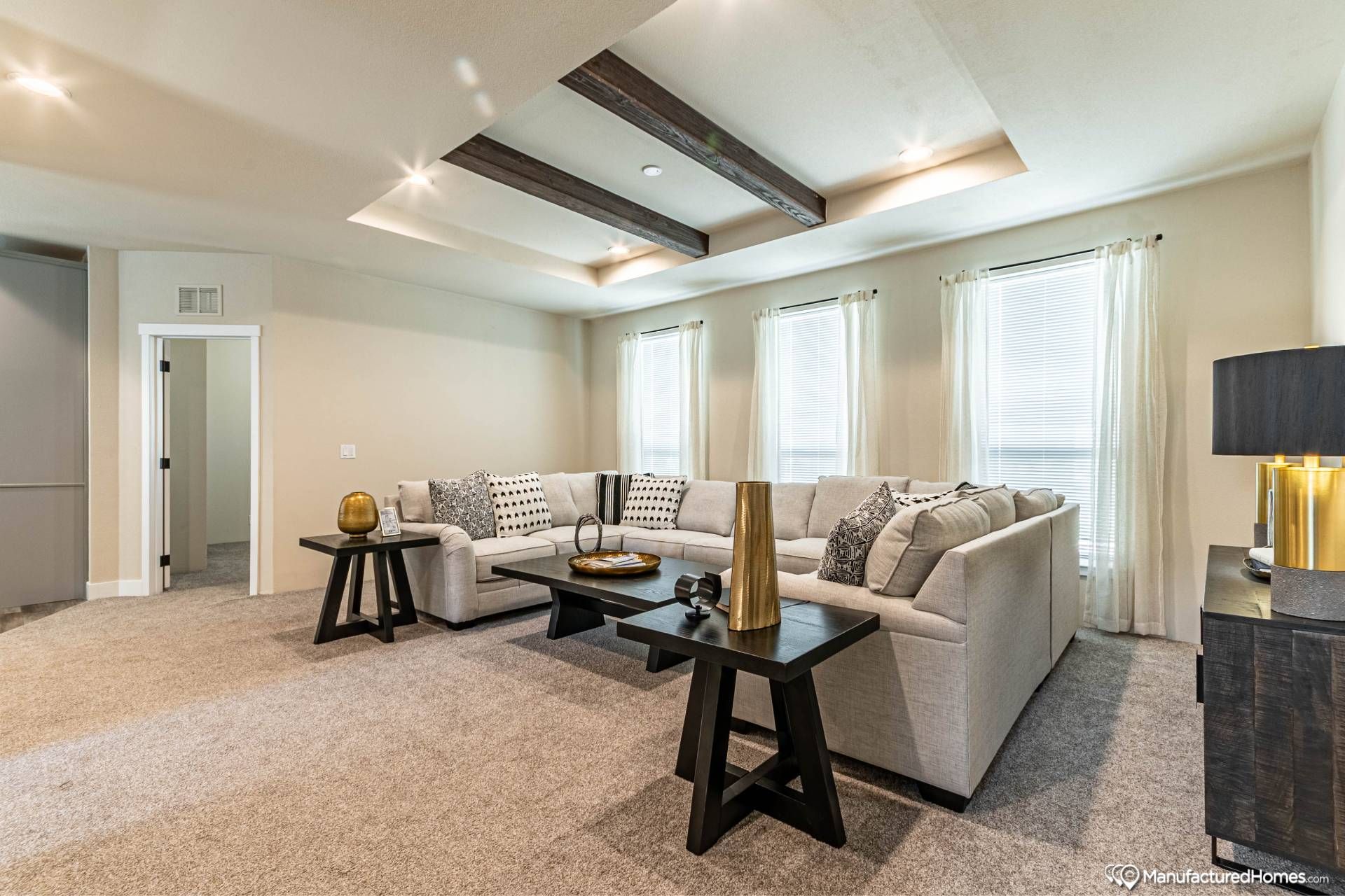 Modern living room with a large beige sectional sofa, black and gold decor accents, a coffee table, and three windows with sheer curtains, conveying a cozy atmosphere.
