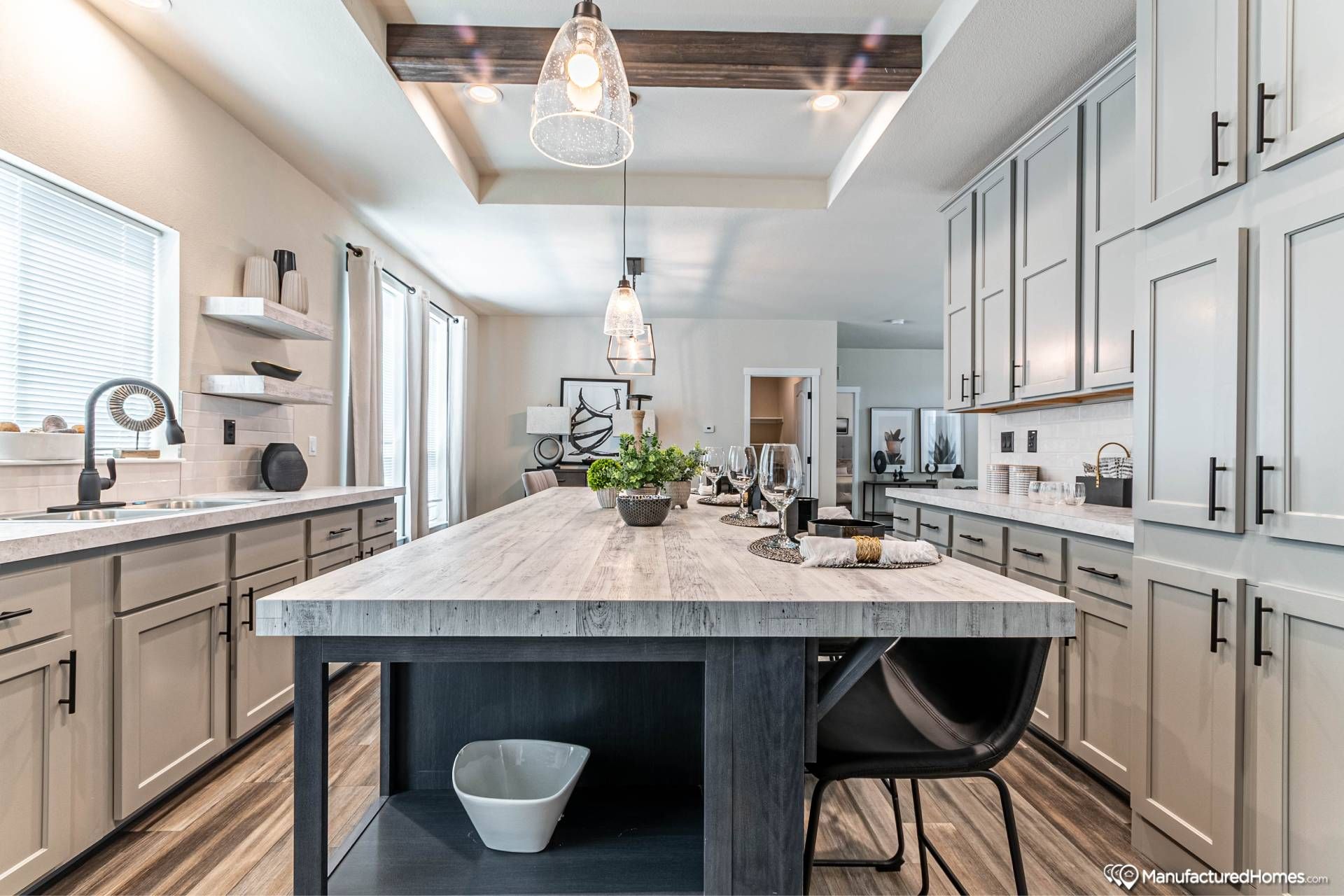 Modern kitchen with a large central island featuring wood countertops and decorative plants. Pendant lights hang above. Cabinets and shelves line the walls.