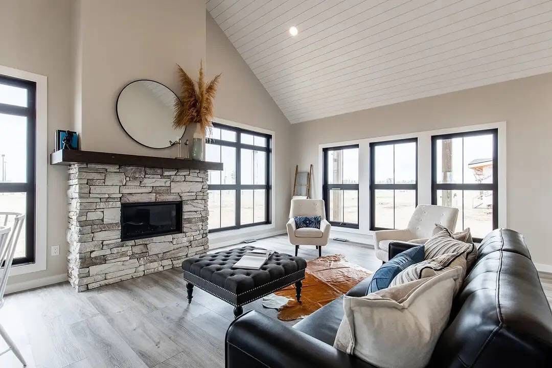 Modern living room with a vaulted ceiling and light gray walls. Features a stone fireplace, large windows, black sofa, and a tufted ottoman. Cozy and bright.