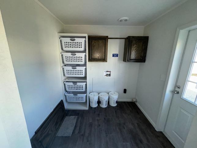 Laundry room with white walls, dark wood cabinets, and a shelf holding five laundry baskets. Three white buckets are under the shelf. The floor is dark wood, and a door is on the right.