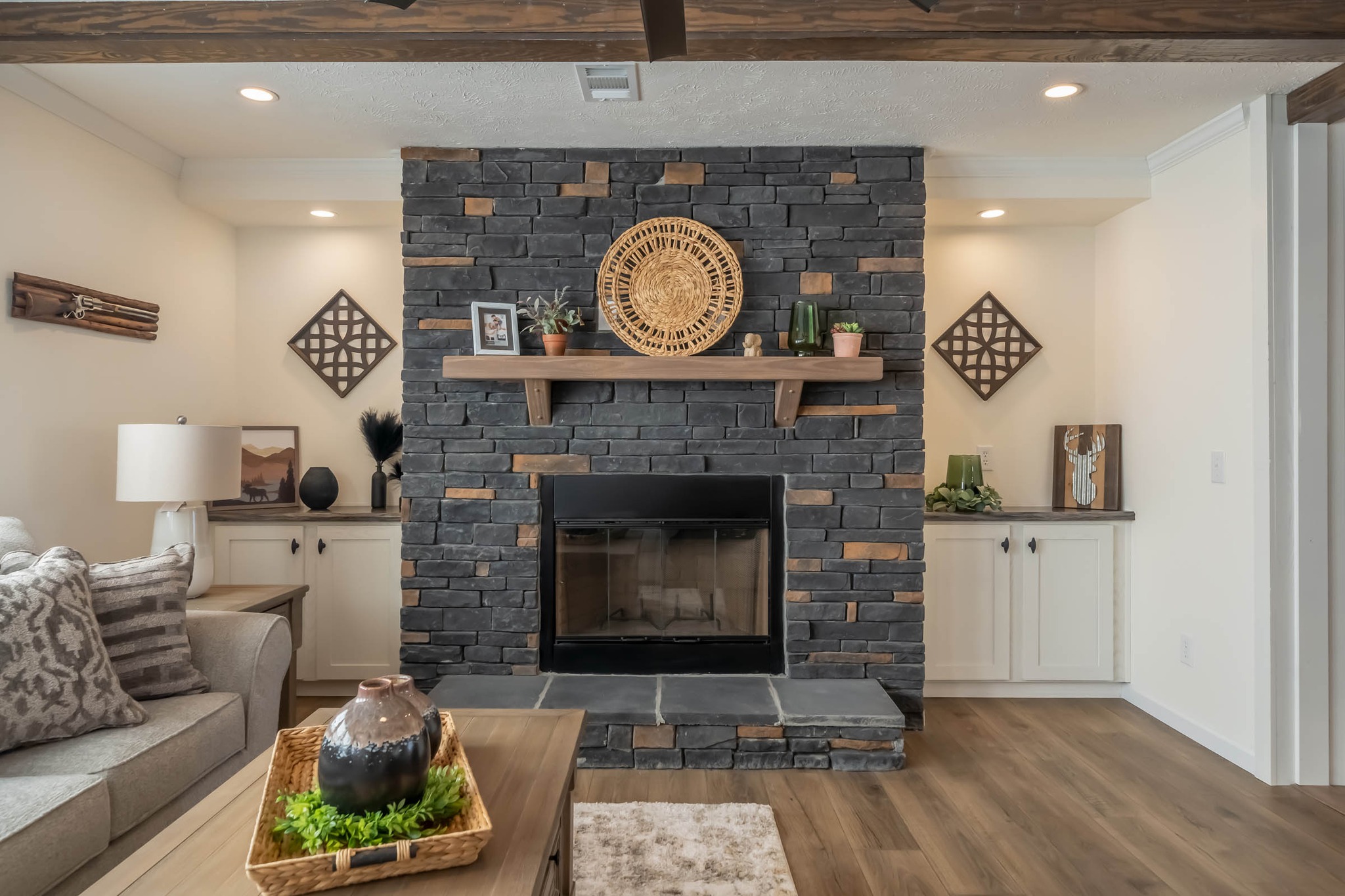 Elegant living room with a dark stone fireplace, wooden mantel, and cozy decor. Neutral tones, plants, and woven art evoke warmth and relaxation.