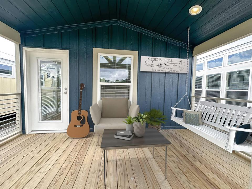 Inviting porch with wooden flooring, blue walls, and a white swing with cushions. A guitar, armchair, and potted plant create a cozy, relaxing vibe.