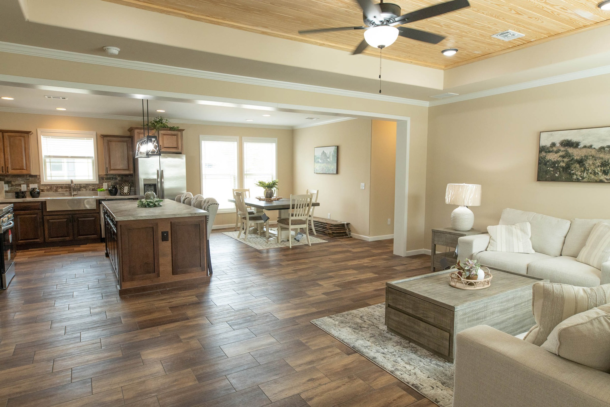 Open-plan living area with a cozy beige couch, wooden coffee table, and a neutral-tone dining table. Kitchen features wood cabinets and a large island. Ceiling fan above enhances the warm, inviting feel.