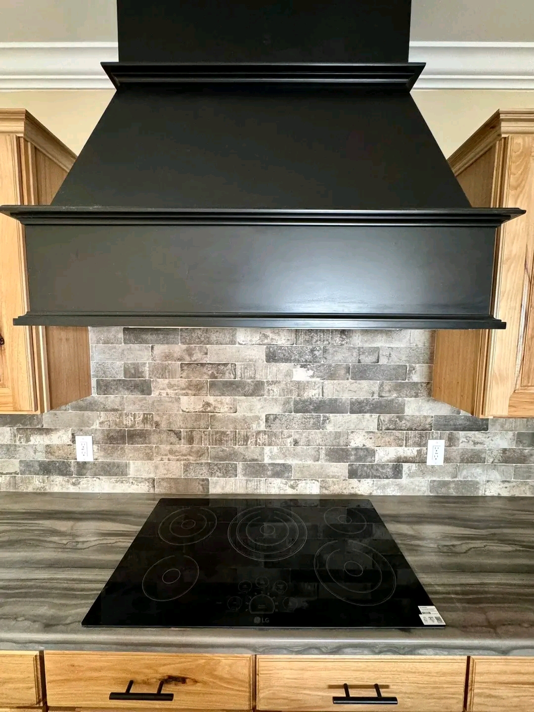 Modern kitchen setup with a large black range hood and electric cooktop. Gray stone backsplash, wooden cabinets, and a sleek countertop.