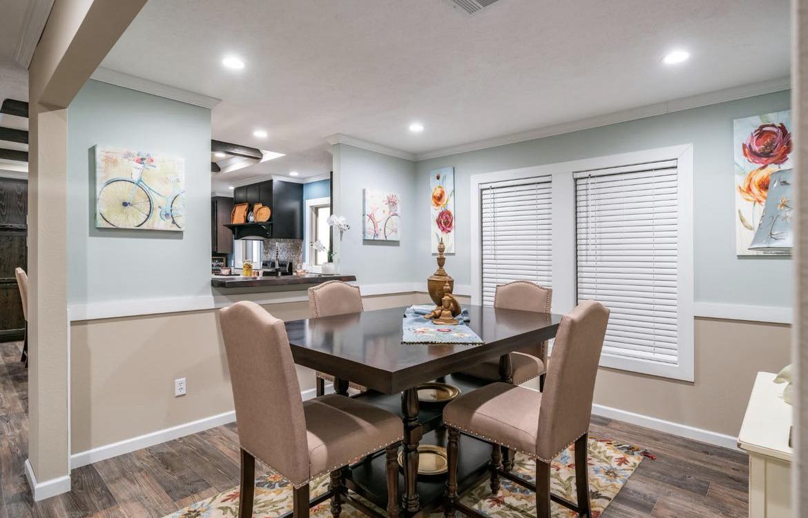 Cozy dining area with a dark wooden table and four beige chairs on a floral rug. Bright art decorates pastel walls, and the space is warmly lit.