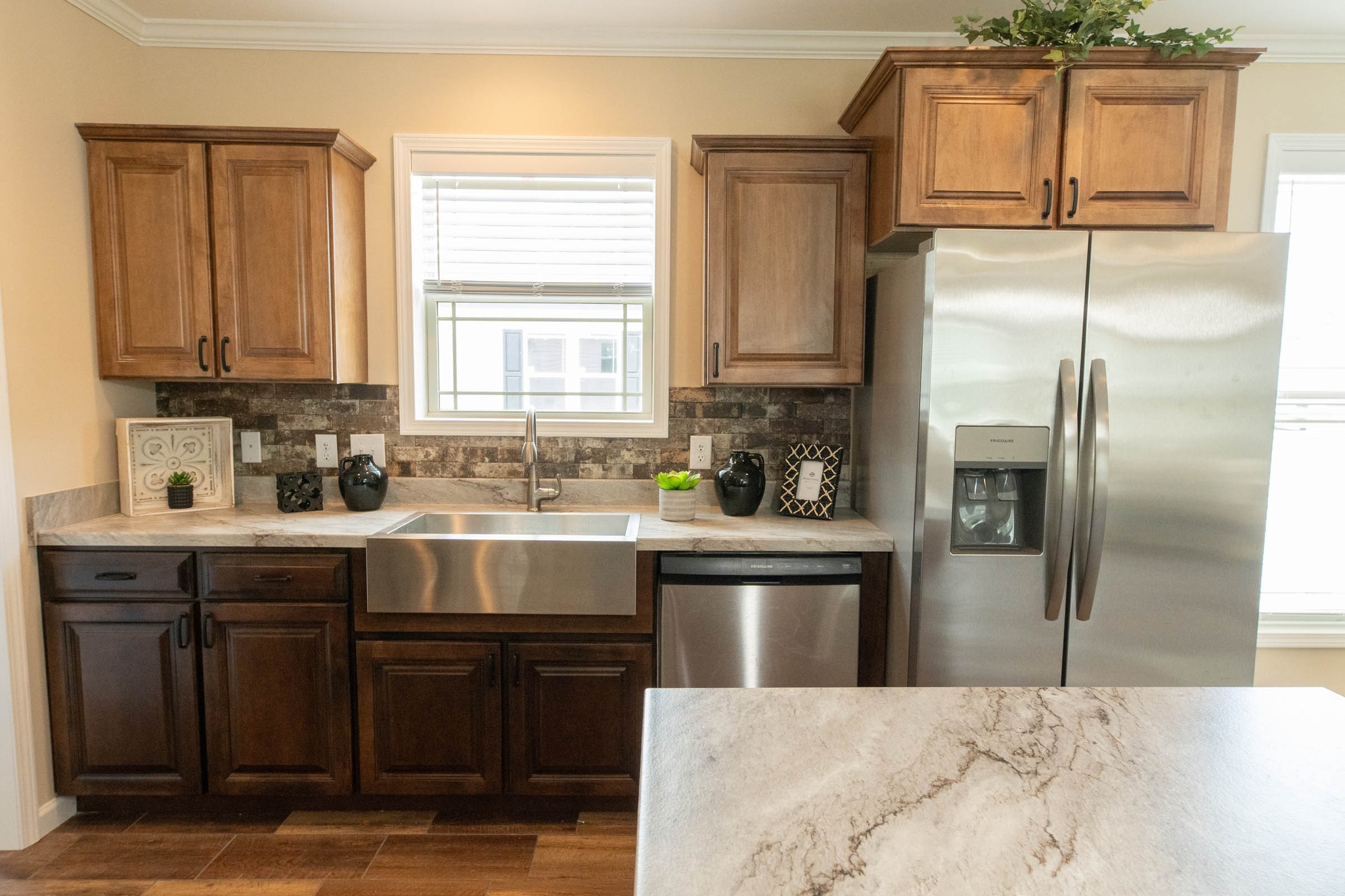 Modern kitchen with wooden cabinets, stainless steel fridge, and farmhouse sink. Marble countertops and decorative plants create a warm, inviting ambiance.