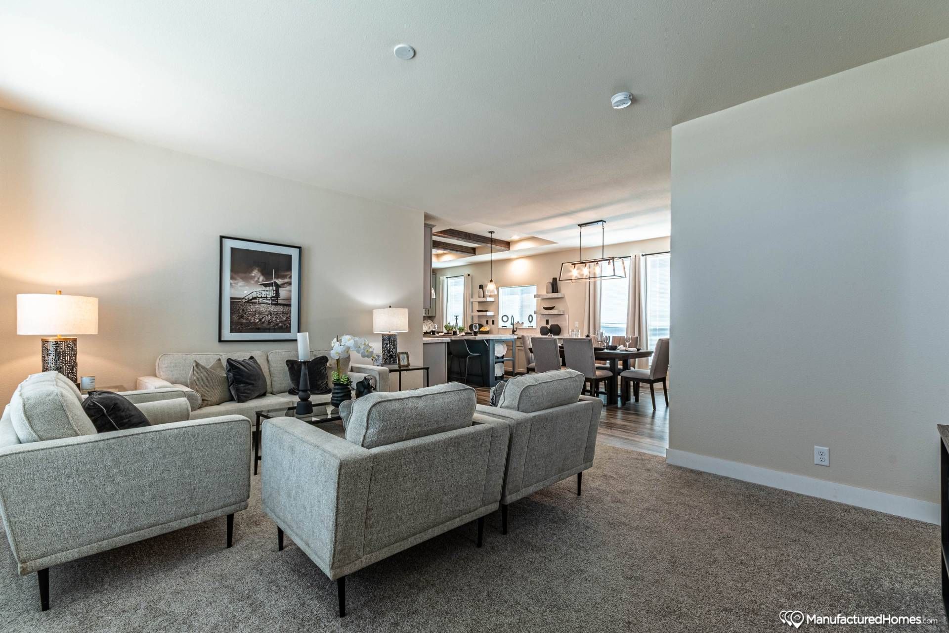 Spacious modern living room with gray sofas, elegant decor, and warm lighting. Open layout leads to a dining area with stylish pendant lights. Cozy and inviting ambiance.