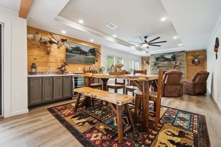 Rustic dining room with wooden table, benches, and vibrant patterned rug. Cozy recliners face a stone fireplace. Mounted animal heads adorn the walls, creating a cabin-like ambiance.