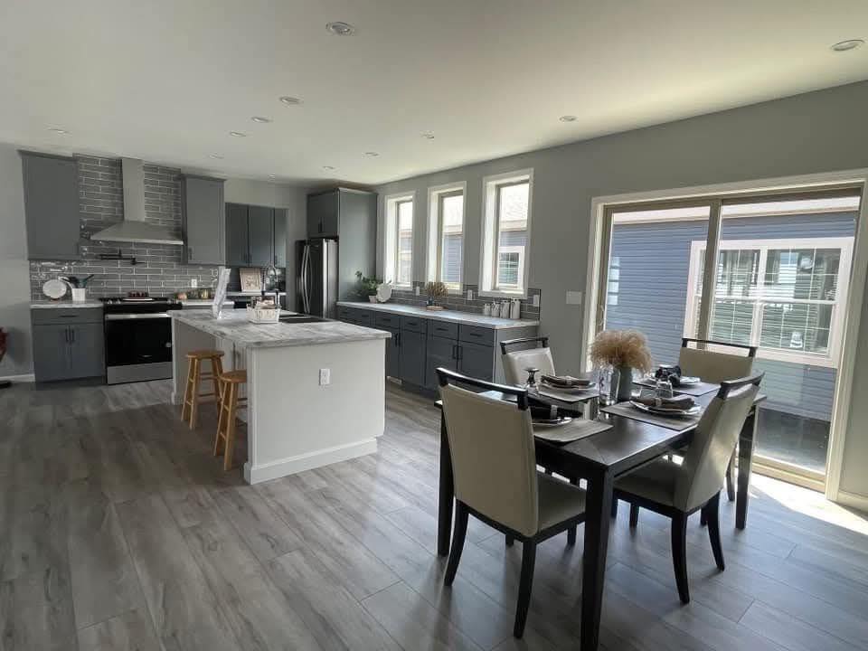 Modern kitchen with light gray cabinets, marble island, and wooden stools. Dining table set for four near large windows. Bright and spacious ambiance.