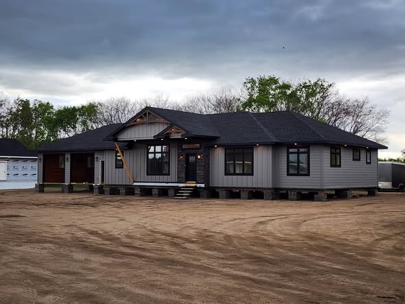 Single-story gray house under construction on stilts, with dark roof, surrounded by dirt, against a cloudy sky. Sparse green trees in background. Calm mood.