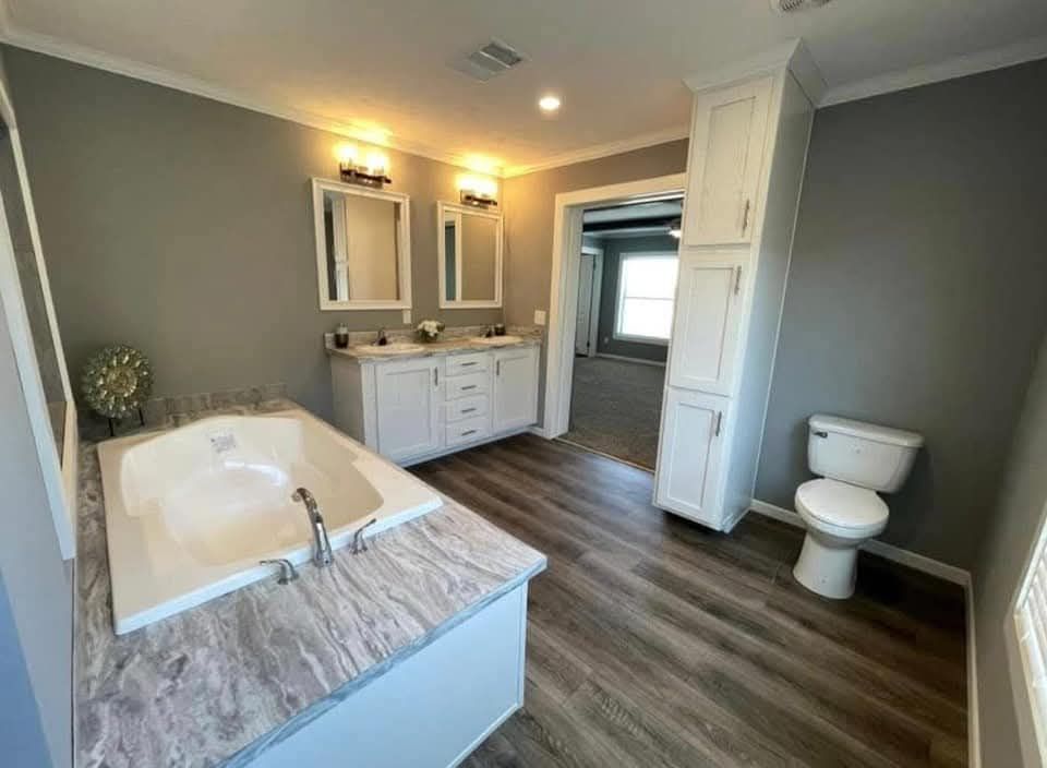 Spacious bathroom with gray walls, featuring a marble-topped bathtub, dual-sink vanity with mirrors, and wood flooring. The atmosphere is modern and clean.
