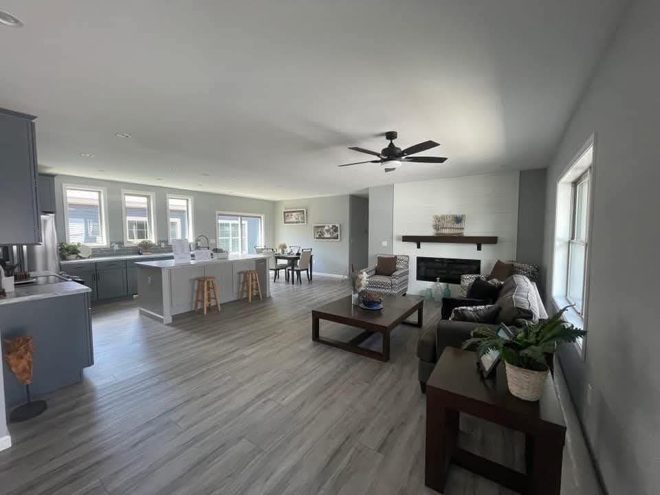 Modern open-concept living room and kitchen in neutral tones, featuring a ceiling fan, cozy sofas, large windows, a fireplace, and a spacious island.