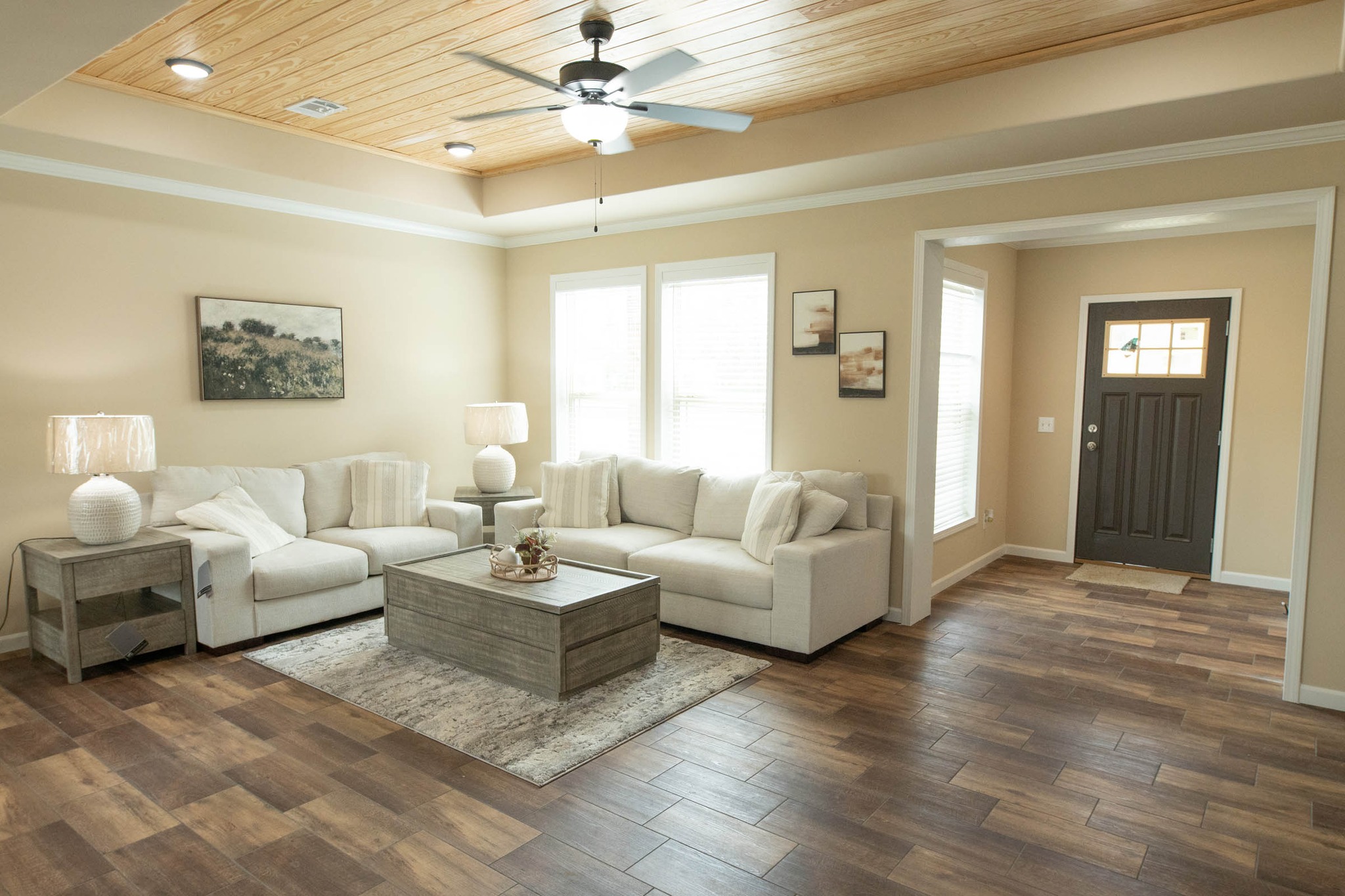 Spacious living room with a beige palette, featuring a large sectional sofa, modern wooden coffee table, and ceiling fan. Natural light from windows creates a cozy ambiance.