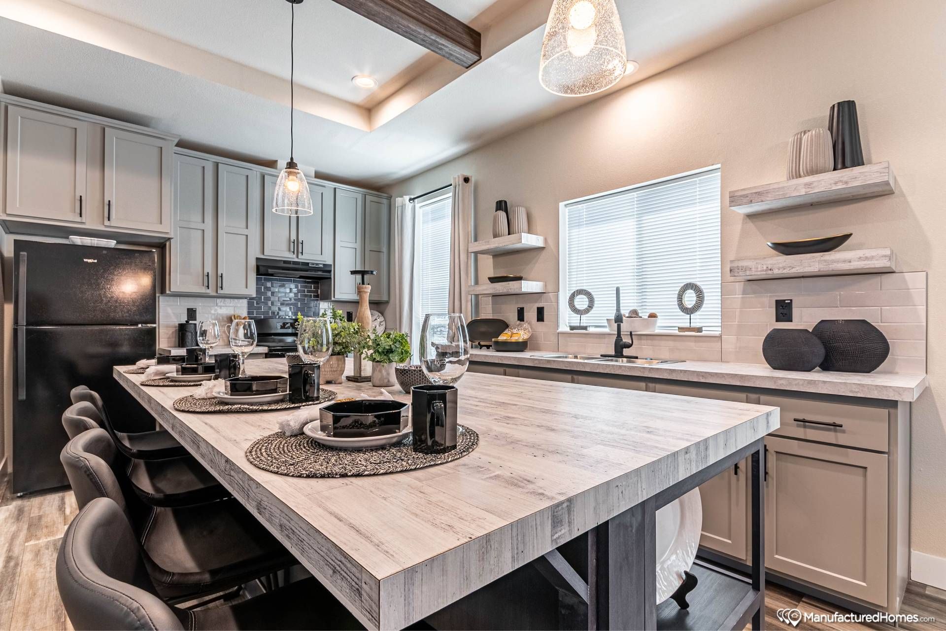 Modern kitchen with a large wooden island, set for dining with black plates and glasses. Grey cabinets, black fridge, decorative shelves, and warm lighting. Cozy and sophisticated ambiance.