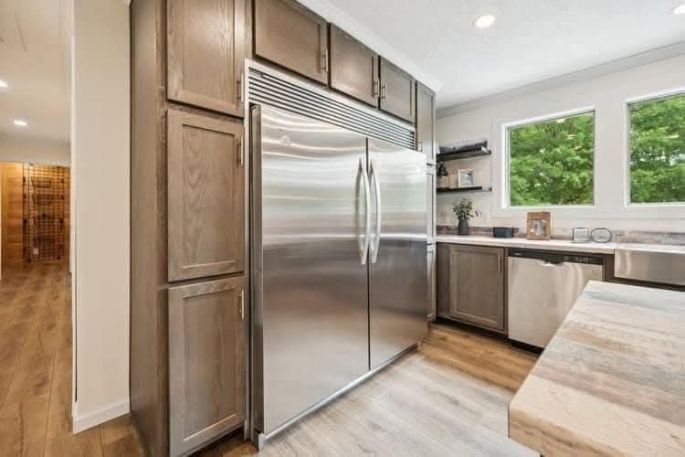 Modern kitchen with a large stainless steel fridge, wooden cabinets, and light-colored countertops. Two windows show green foliage outside.