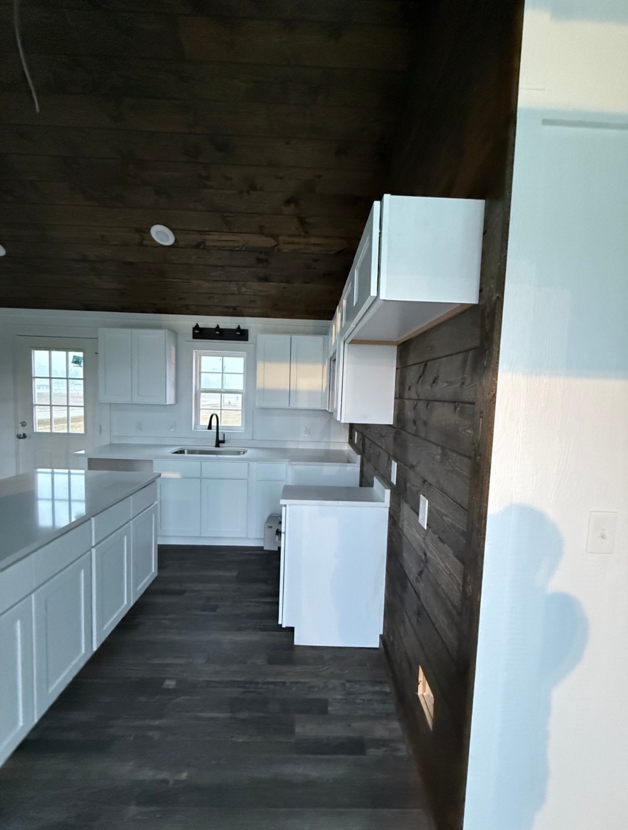 Modern kitchen with white cabinets and dark countertops. Wooden ceiling and wall add warmth. A window above the sink brings in natural light.