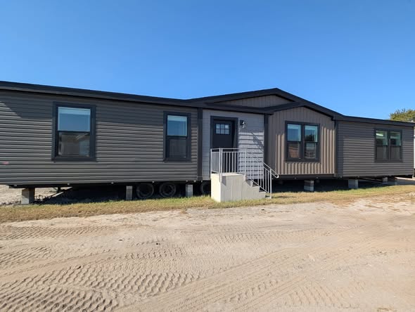 A brown manufactured home with three sections, mounted on wheels, sits on a dirt lot under a clear blue sky, giving a sense of simplicity and functionality.