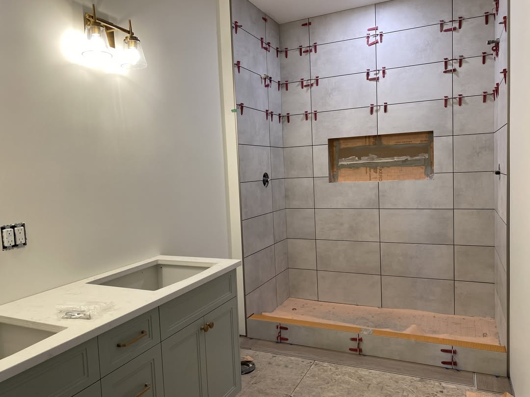 Bathroom under renovation with gray tiles being installed in a shower area. A double vanity with a marble countertop and brass faucet sits nearby. Bright lighting creates a clean, modern feel.
