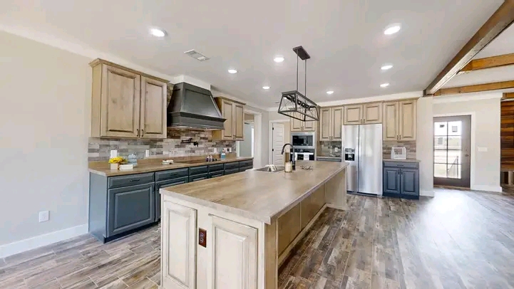 Spacious kitchen with rustic wood flooring, a large island with a faucet, light wood cabinetry, stainless steel appliances, and modern lighting.