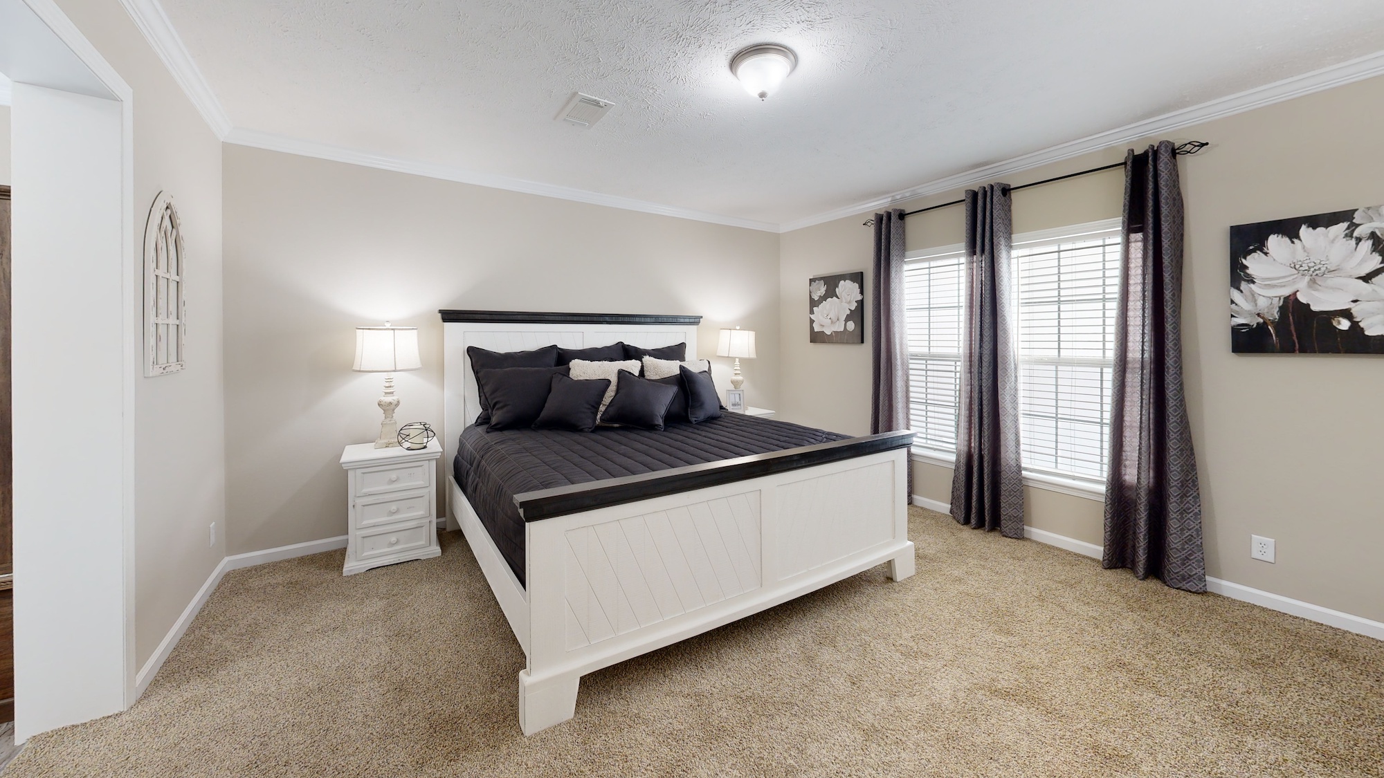 A cozy bedroom with beige walls features a large white bed with dark bedding. Two bedside lamps softly illuminate the space. A window with heavy curtains and a floral picture add a serene touch.