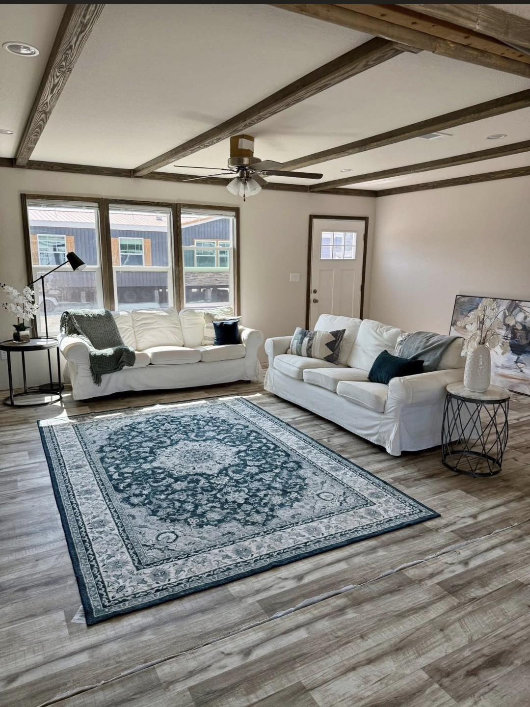 Bright, cozy living room with two white sofas on a wood floor. A teal-patterned rug centers the space. Large windows let in natural light, creating an inviting atmosphere.