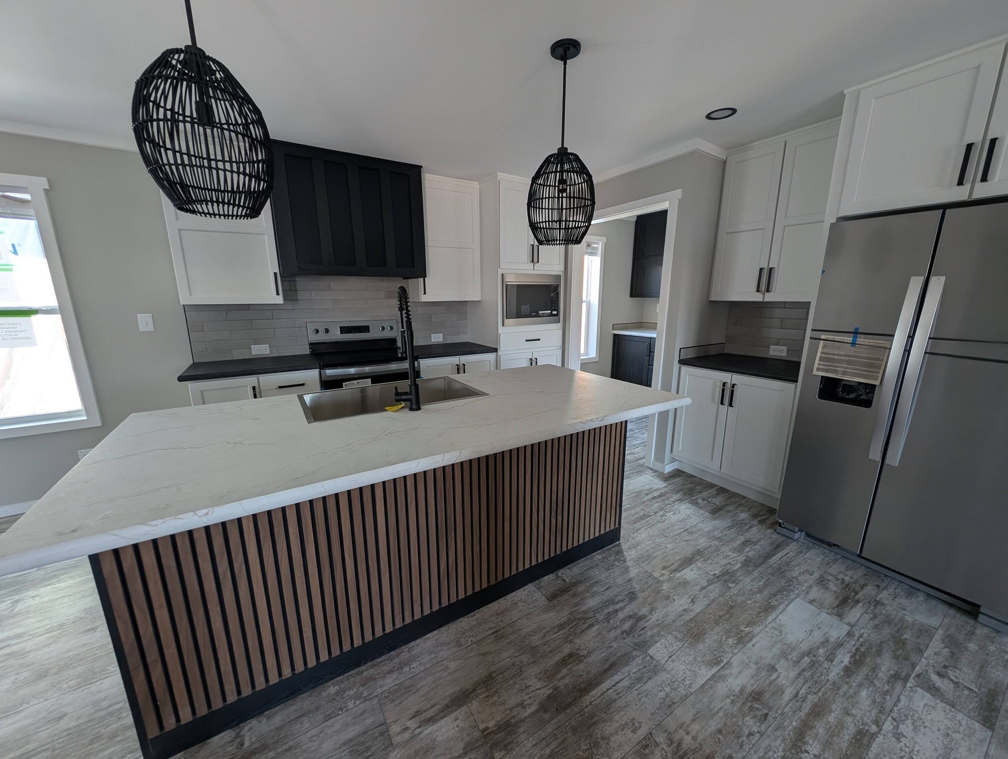 Modern kitchen with wood-paneled island, marble countertop, black fixtures. White and black cabinets, stainless steel fridge, and pendant lights.