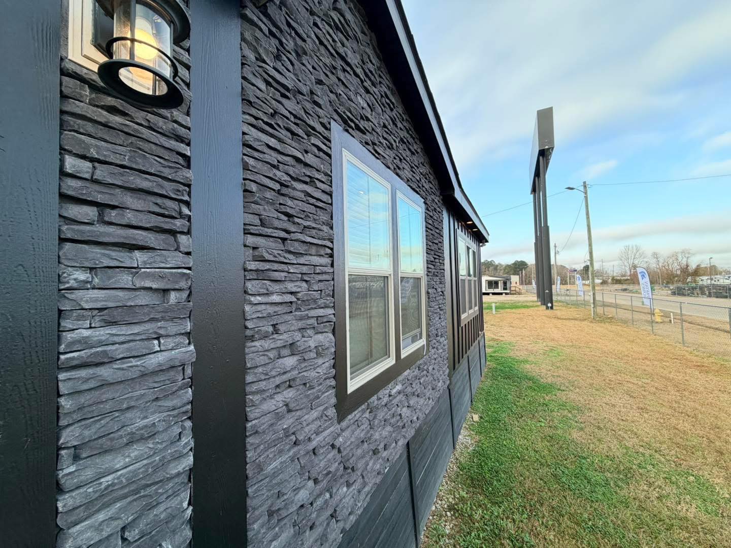 Textured stone wall of a modern building with framed windows. A lamp is mounted on the wall. A grassy area and street are visible under a blue sky.