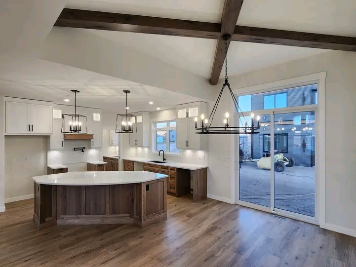 Spacious kitchen with a large island, white countertops, wooden cabinets, exposed beams, modern hanging lights, and large sliding glass doors.