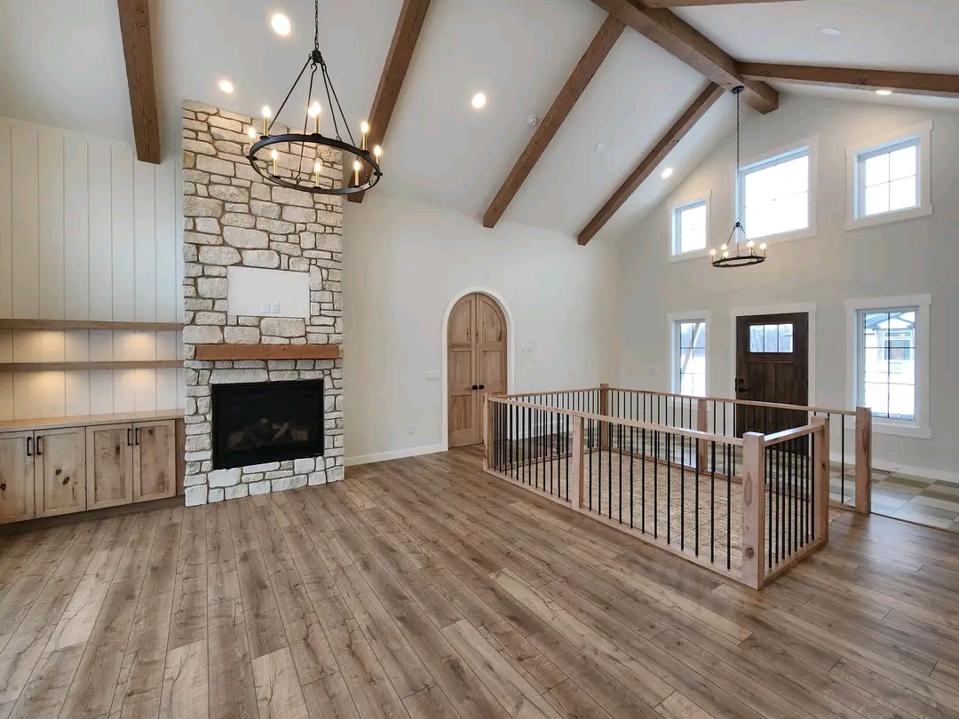 Bright, spacious living room with high vaulted ceiling and wooden beams. Features stone fireplace, rustic chandelier, wood floors, and large windows.