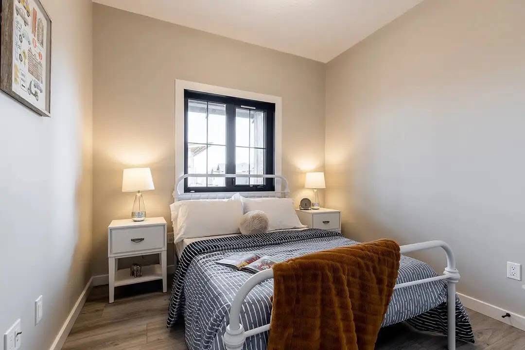A cozy bedroom with beige walls features a double bed with a striped duvet, white pillows, and a fluffy cushion, flanked by two white nightstands with lit lamps. A brown throw is draped over the bed, creating a warm, inviting atmosphere. A window with black trim is above the bed, and a framed artwork hangs on the left wall.